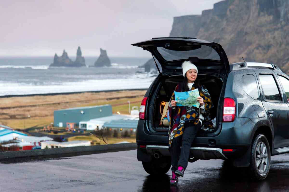 Traveler sitting in the open trunk with a map, parked by the sea and cliffs on a rainy day.