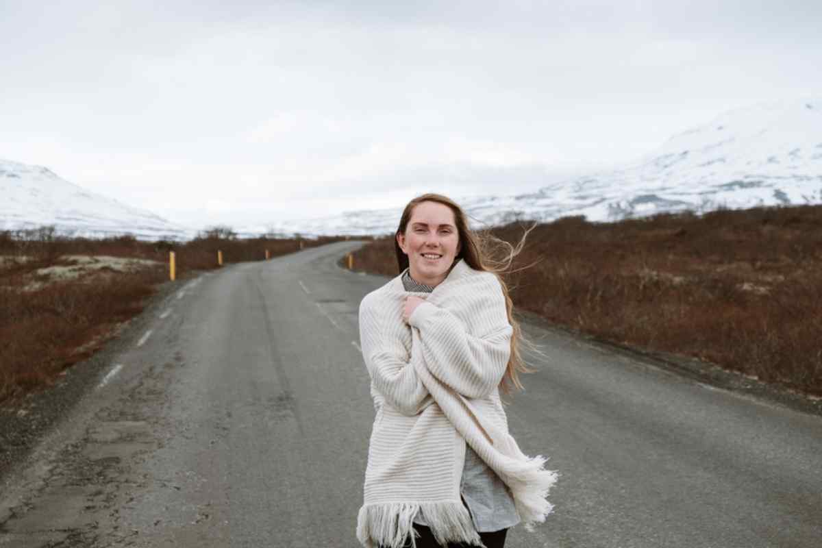 Safety in Iceland Smiling traveler wrapped in a warm blanket standing on an empty Icelandic road, with brown tundra and snow-covered mountains in the background.