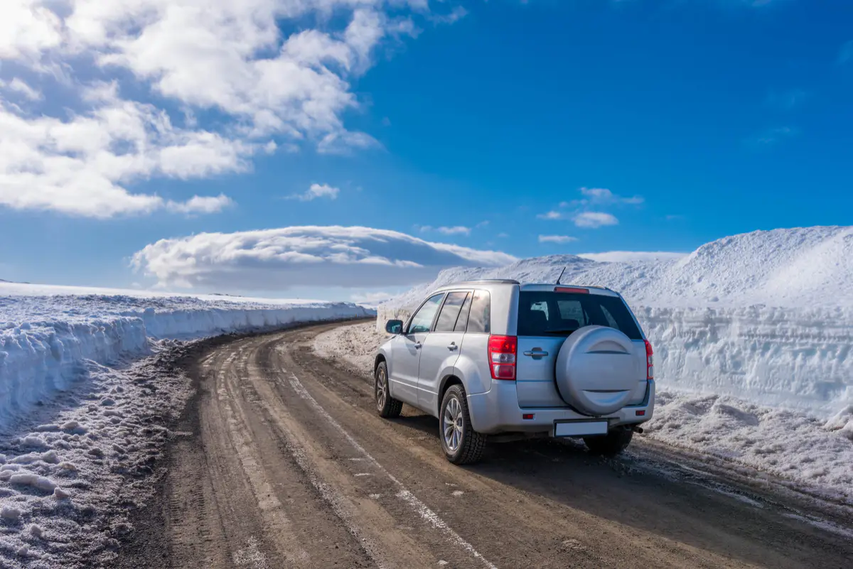 A silver 4x4 SUV driving on a partially snow-covered gravel road in Iceland, surrounded by high snowbanks under a bright blue sky.