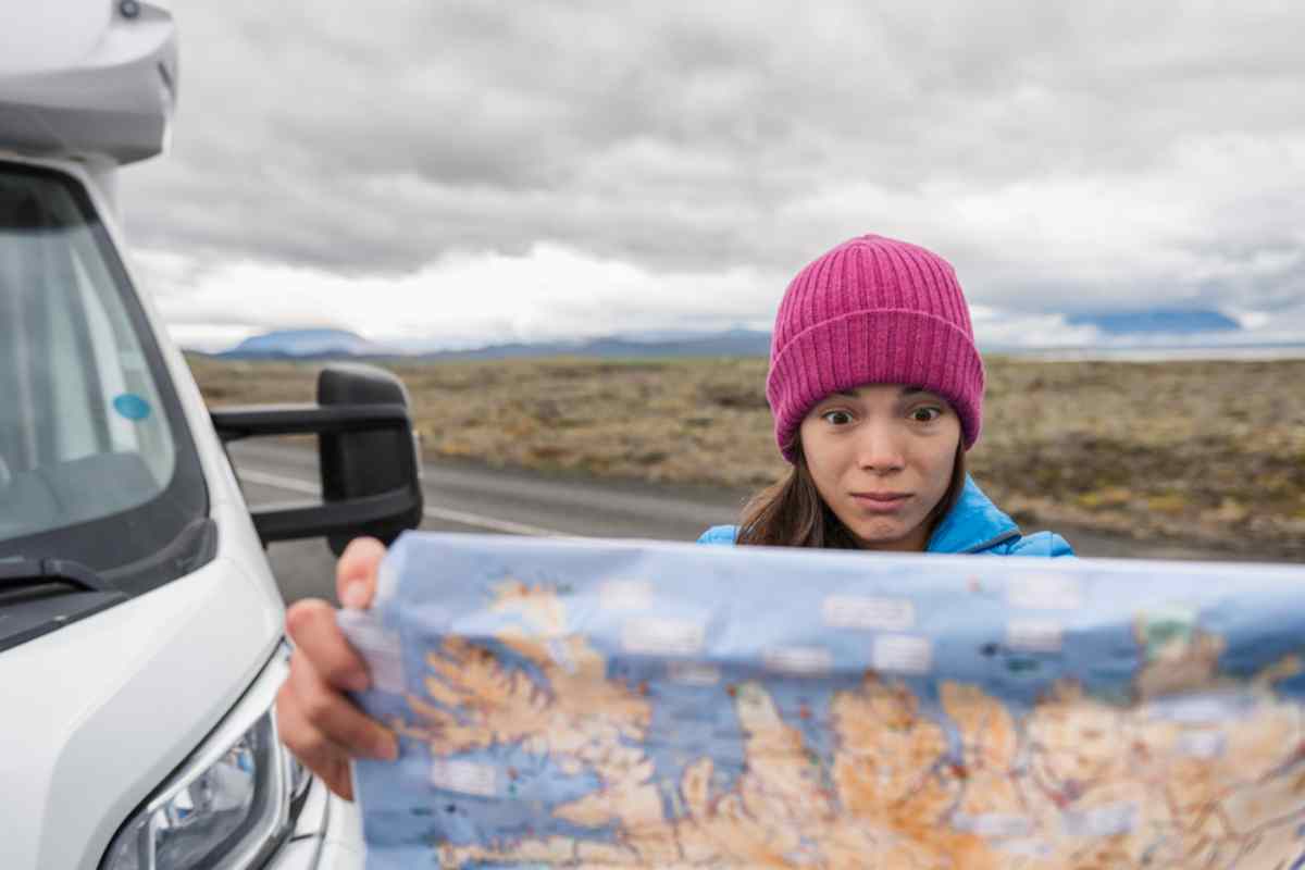A sheltered woman with a worried face looks at a physical map in her hands as she stands on the road outside her camper.