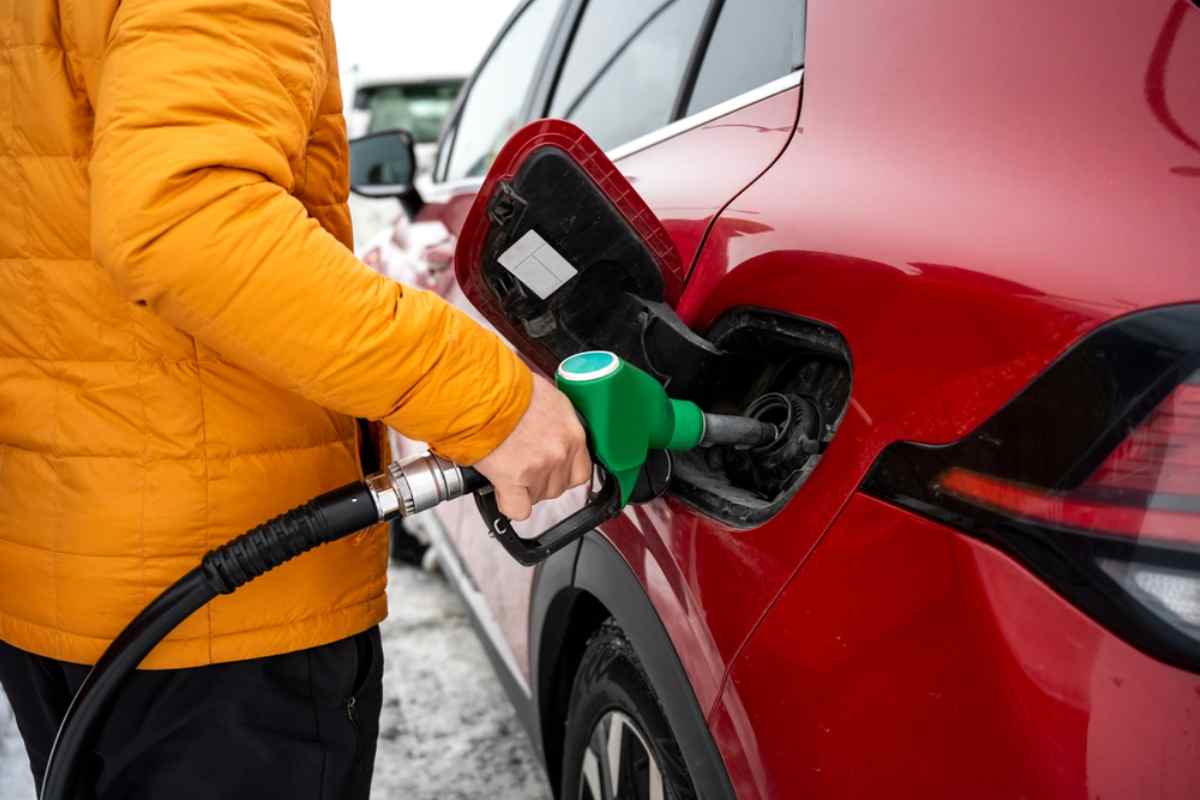 A person refuels a red car at an Icelandic gas station by grabbing the gas pump with one of his hands and inserting it into the vehicle's tank.
