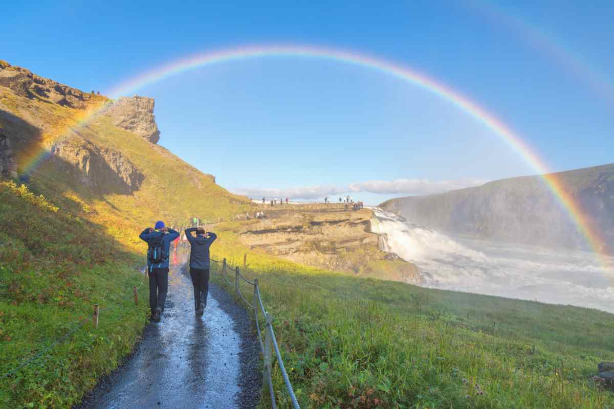 General view of a trail near the Gullfoss waterfall in Iceland, where two tourists are walking in hyper waterproof clothing with a beautiful rainbow above them, detached from the group.