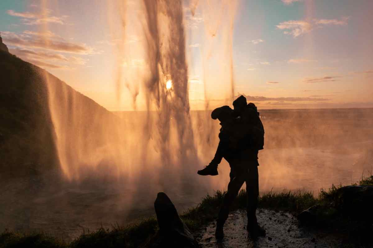 A couple kiss as one person stands slightly above the other in the shade with the sun beating down on Iceland's Seljalandsfoss waterfall in front of them.