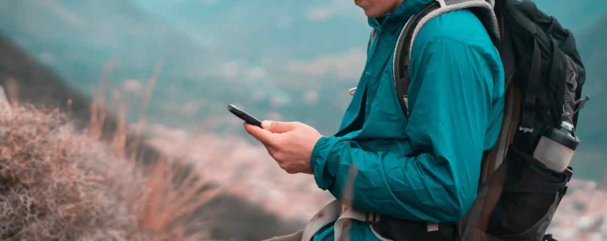 A hiker in hiking clothes clutches a cell phone in his hand as he stands outdoors.