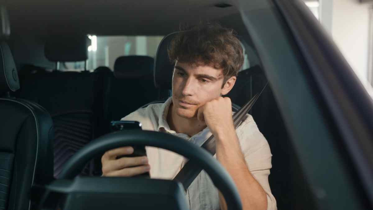 A young man from the driver's seat of a parked vehicle looks listlessly at his cell phone.