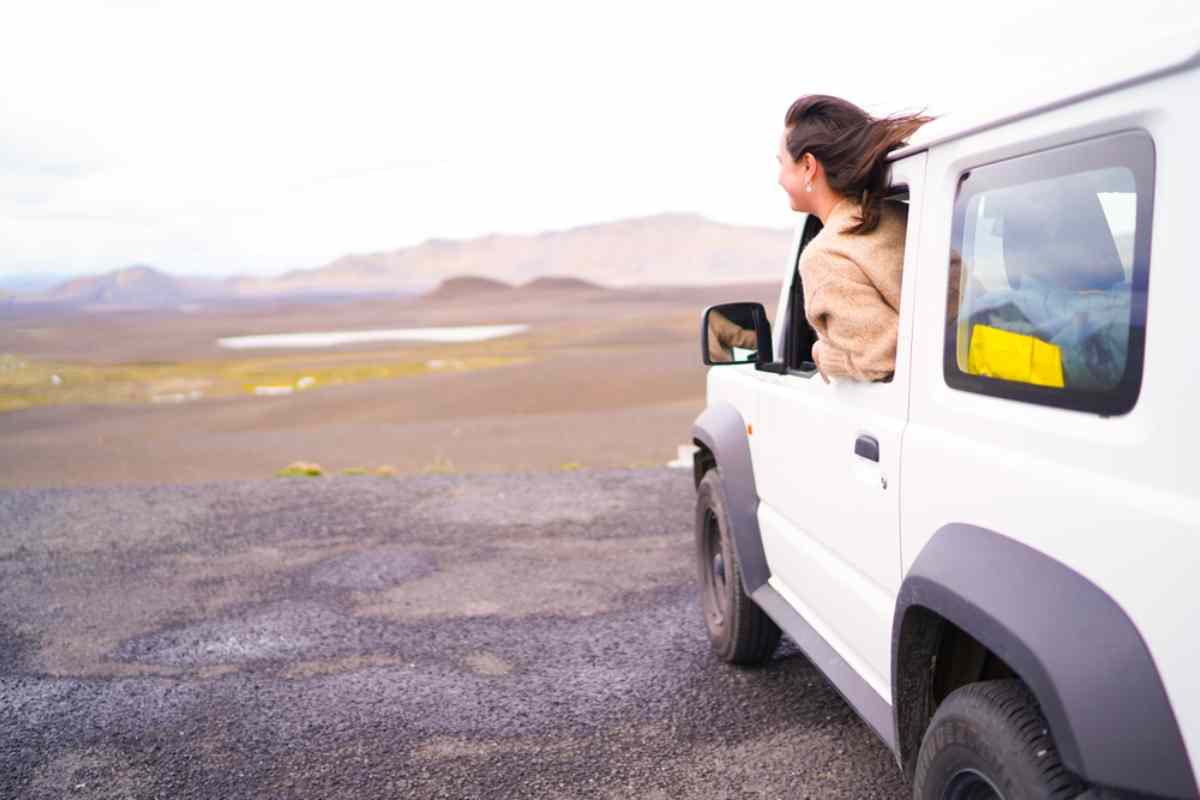 A woman sticks her head out of the window of a moving 4x4 as it travels through an arid, unpaved island landscape.