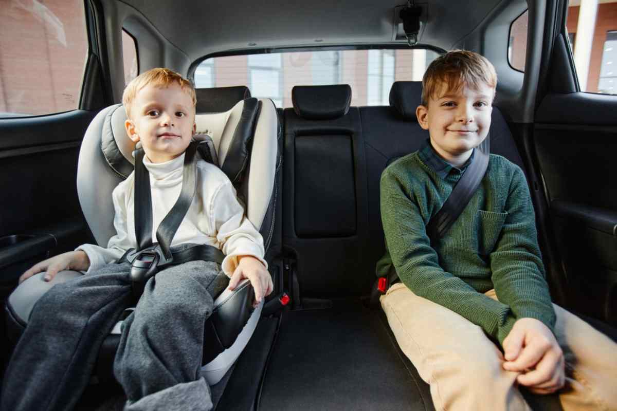 Two children of different ages remain seated in the back seat of a car, one with a car seat and one without, but both wearing a seat belt.