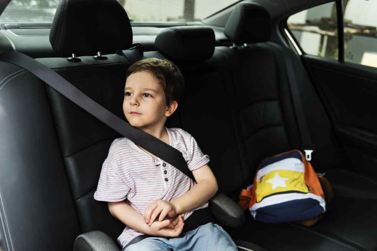 A small child is sitting with a seat belt and booster in the back seat of a parked car.
