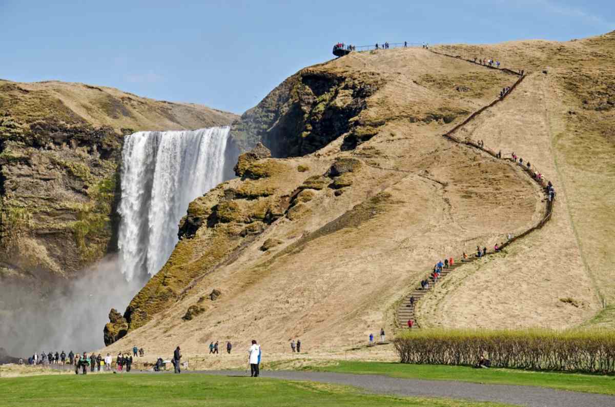 Skógafoss waterfall in Iceland with tourists hiking up the trail alongside the cliff Climbing the stairs at Skogafoss waterfall