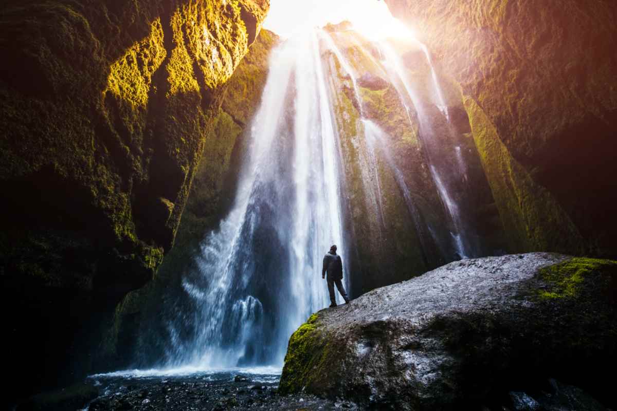 Gljufrabui Waterfall Traveler on a boulder inside a mossy slot canyon, gazing up at Gljufrabui waterfall near Seljalandsfoss as sunbeams pour through the opening.