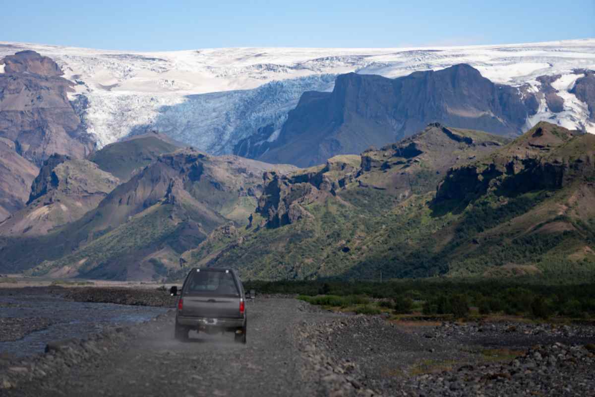 Safety tips for the F249 road in Iceland 4x4 driving the F249 gravel road toward Thorsmork, dust trailing beside a braided river, with steep green cliffs and glacier-capped peaks ahead.