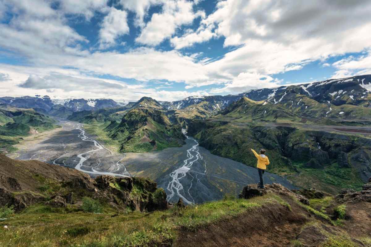 Thorsmork Nature Reserve Panoramic view over Thorsmork valley: braided rivers, rugged green mountains with snow patches, and a hiker in a yellow jacket standing on the rim.