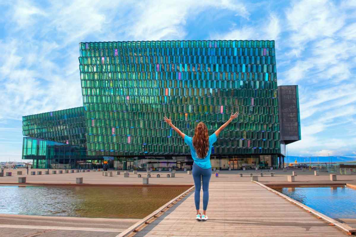 Harpa Concert Hall in Reykjavik with its glass fa&ccedil;ade, reflected colors, and a visitor standing on the waterfront walkway.